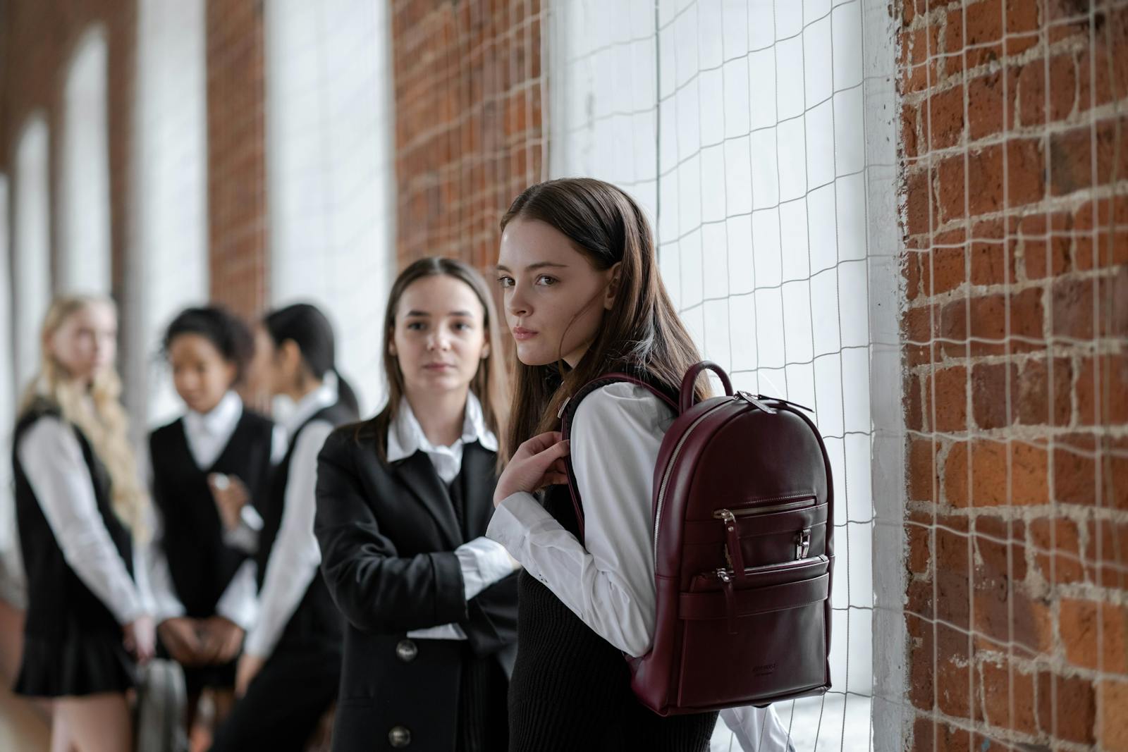 Five schoolgirls in uniforms standing in a brick hallway, carrying backpacks.
