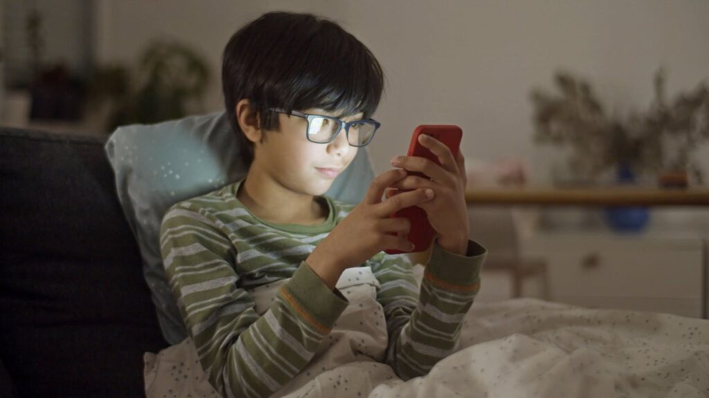A young boy with eyeglasses using a smartphone in bed at night.