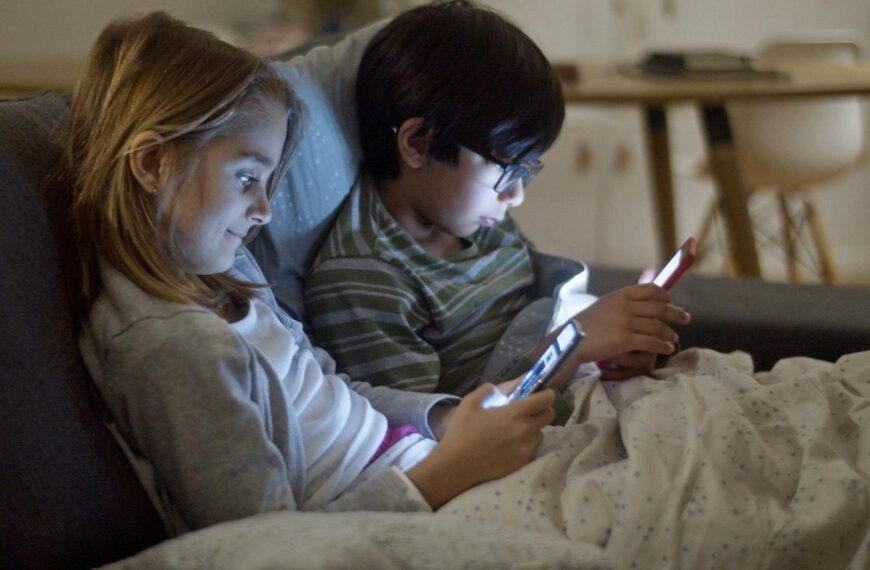 Two children using smartphones at night, indoors, under blankets.