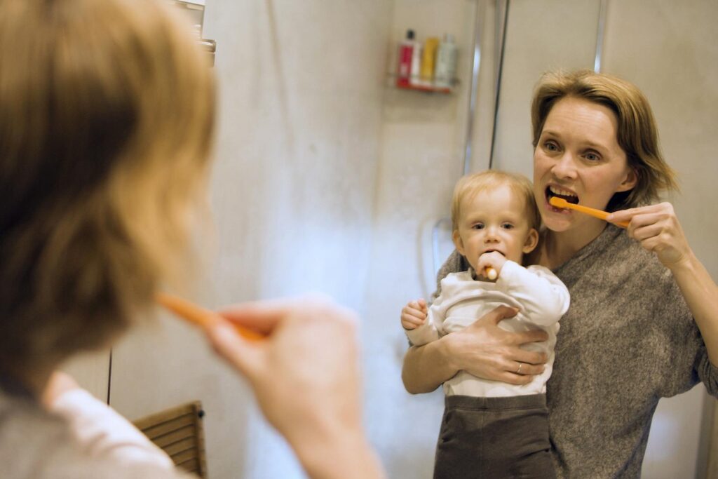 A mother and her young child brushing teeth together in a bathroom mirror, showcasing daily hygiene routine.