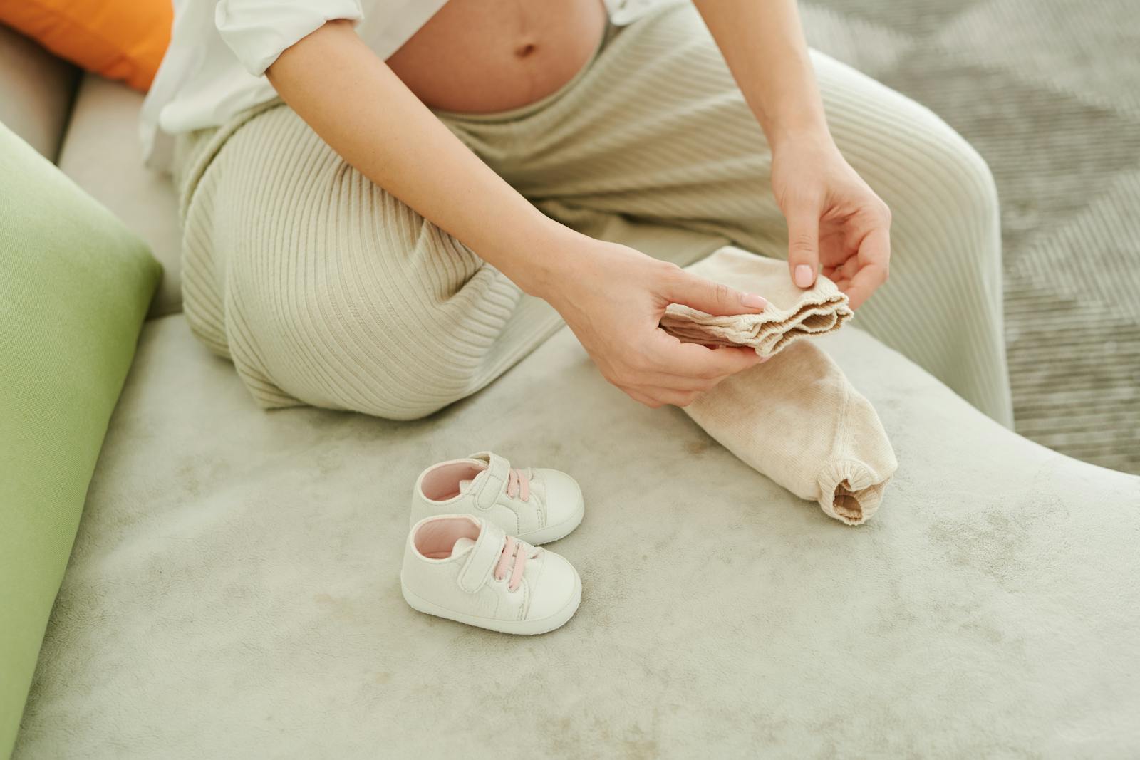 Pregnant woman folding baby clothes with baby shoes on the couch.