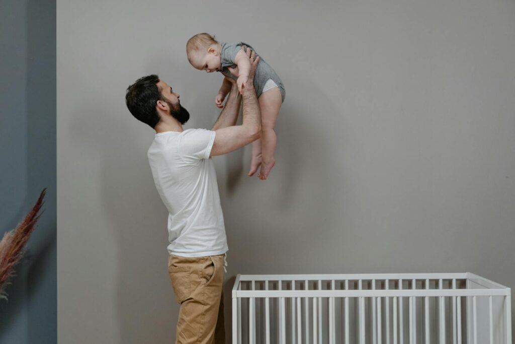 A father joyfully lifts his baby near a crib, creating a moment of bonding and happiness in a nursery setting.