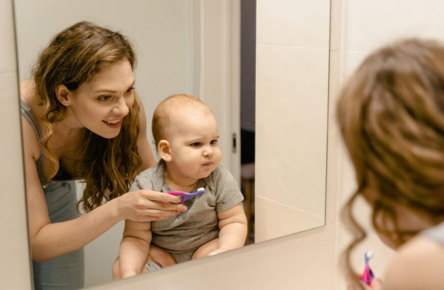 A mother helps her baby brush teeth while they share a joyful moment in front of a bathroom mirror.