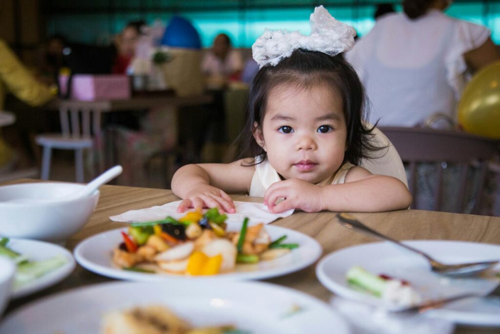 Cute toddler girl at a restaurant table surrounded by various dishes, enjoying her meal