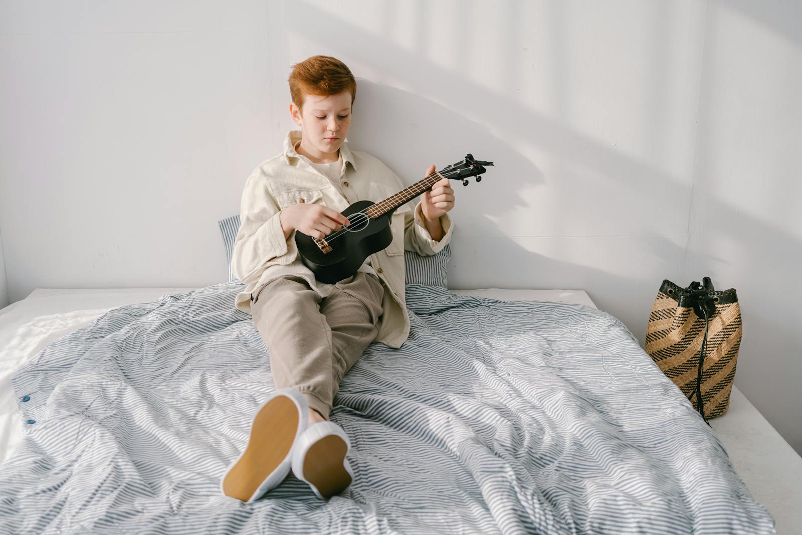 Young boy with red hair playing a ukulele while sitting on a bed, enjoying a peaceful moment indoors.