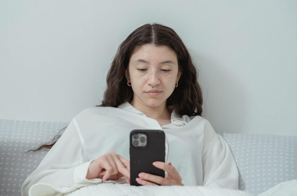 A young woman wearing pajamas relaxes in bed while browsing on her smartphone at home.