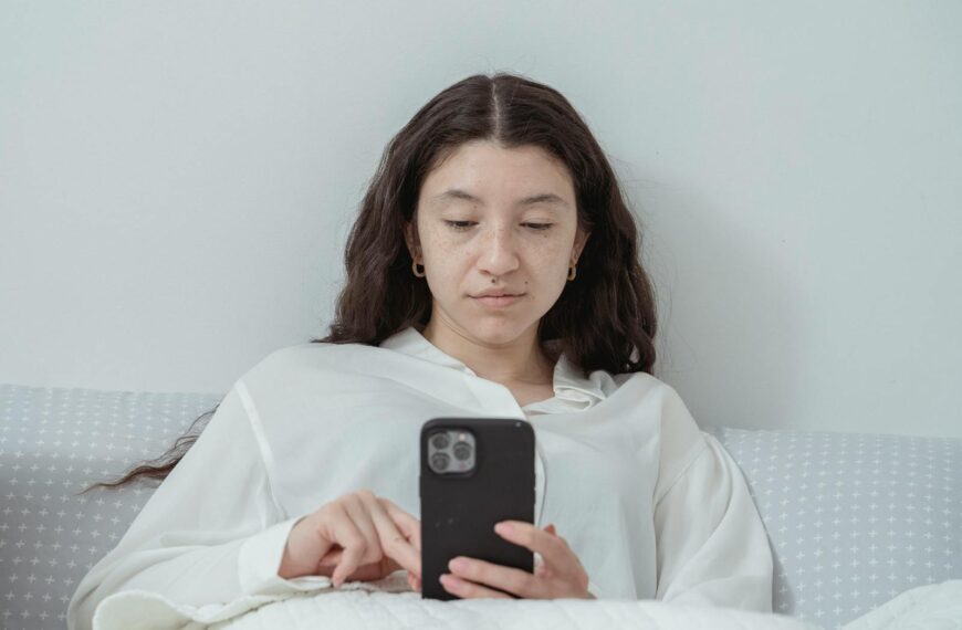 A young woman wearing pajamas relaxes in bed while browsing on her smartphone at home.