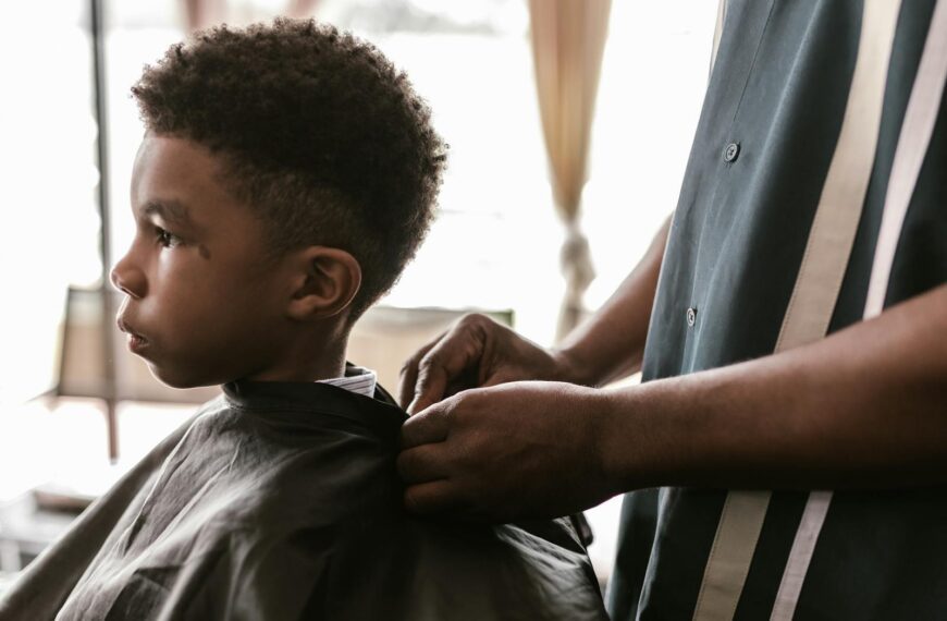 A child receives a haircut in a barbershop, wearing a cape, with a focus on the process.