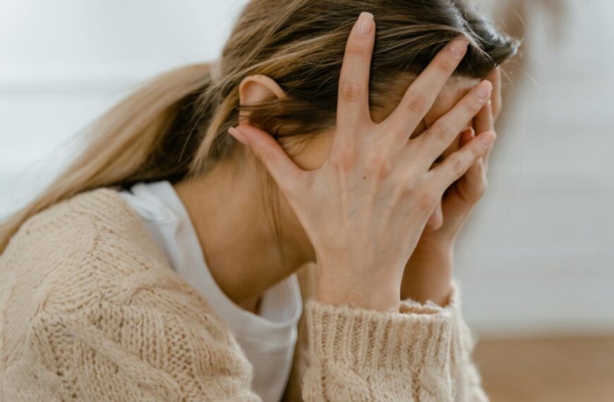 Woman sitting indoors with face covered by hands, expressing stress and frustration.