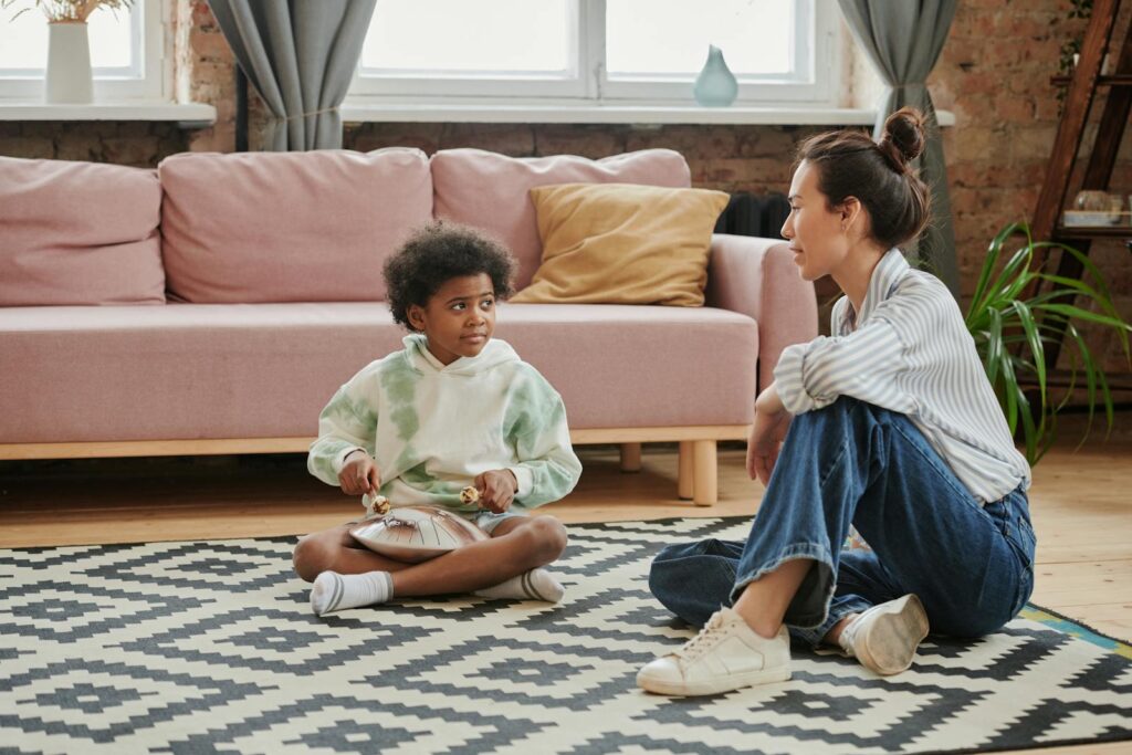 A child playing a steel drum while sitting indoors with a mentor nearby.