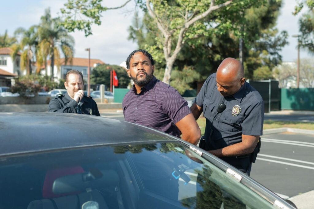 Two police officers detaining a suspect beside a car in a sunny outdoor setting.