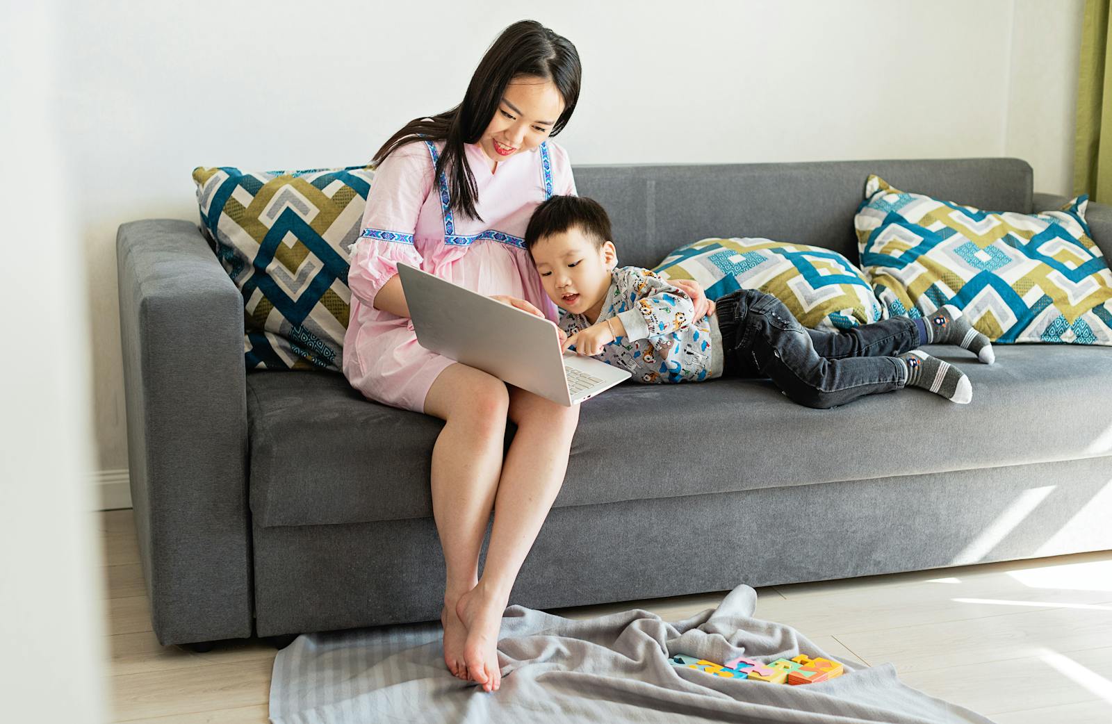 Pregnant mother and son bonding on sofa, using laptop at home.