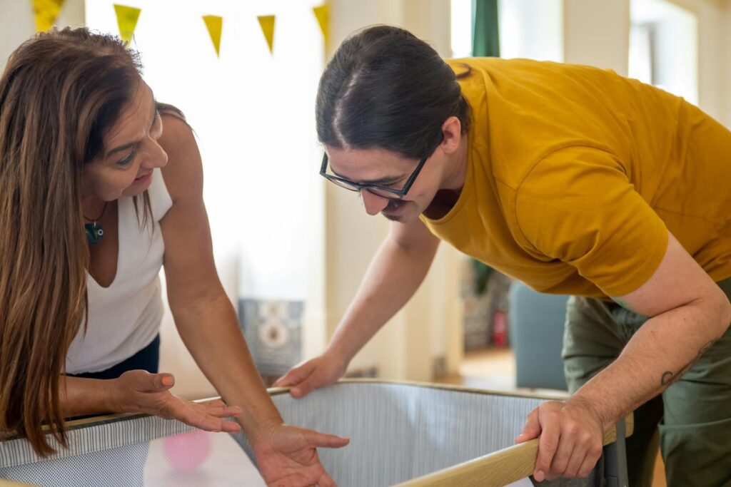 A couple happily assembles a baby crib at home, smiling and engaged in conversation.