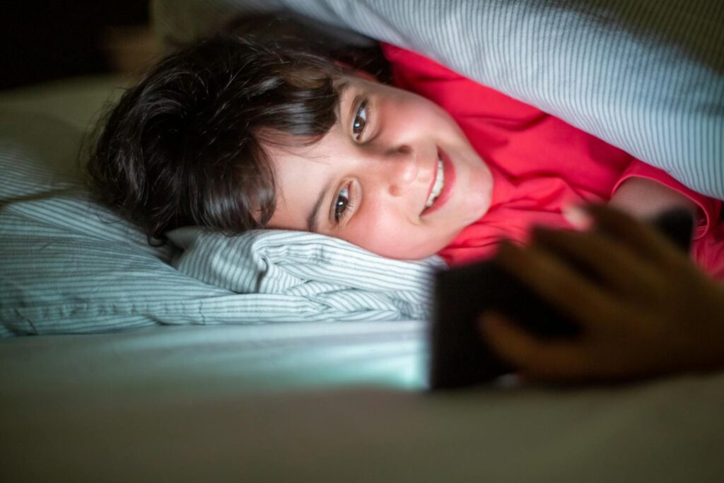 A cheerful young girl smiling at her phone under cozy bed covers.