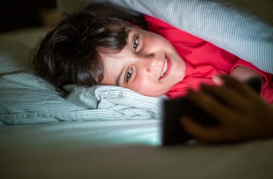A cheerful young girl smiling at her phone under cozy bed covers.