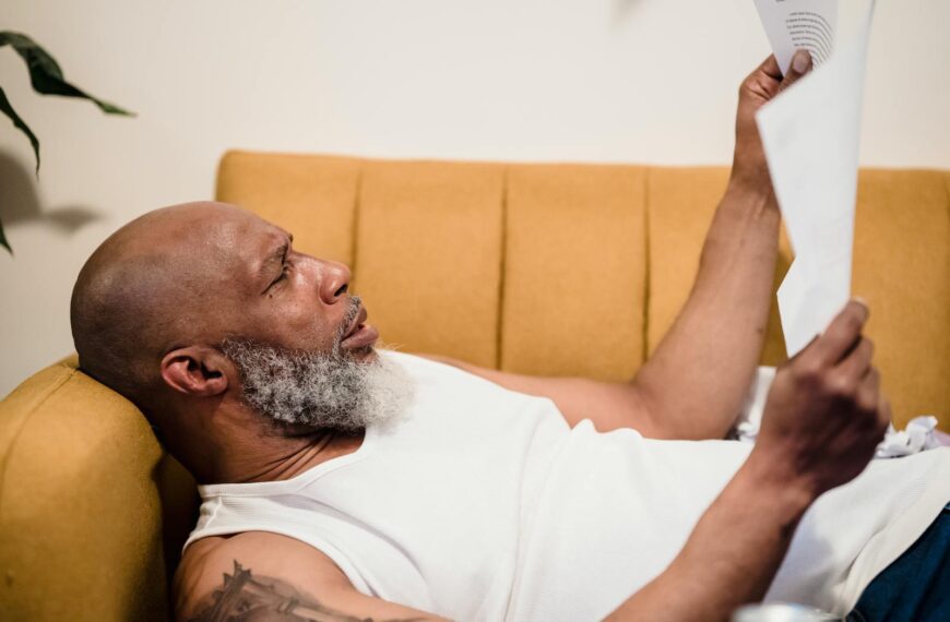 Bald man with facial hair lies on a mustard couch, reading documents in a relaxed setting.