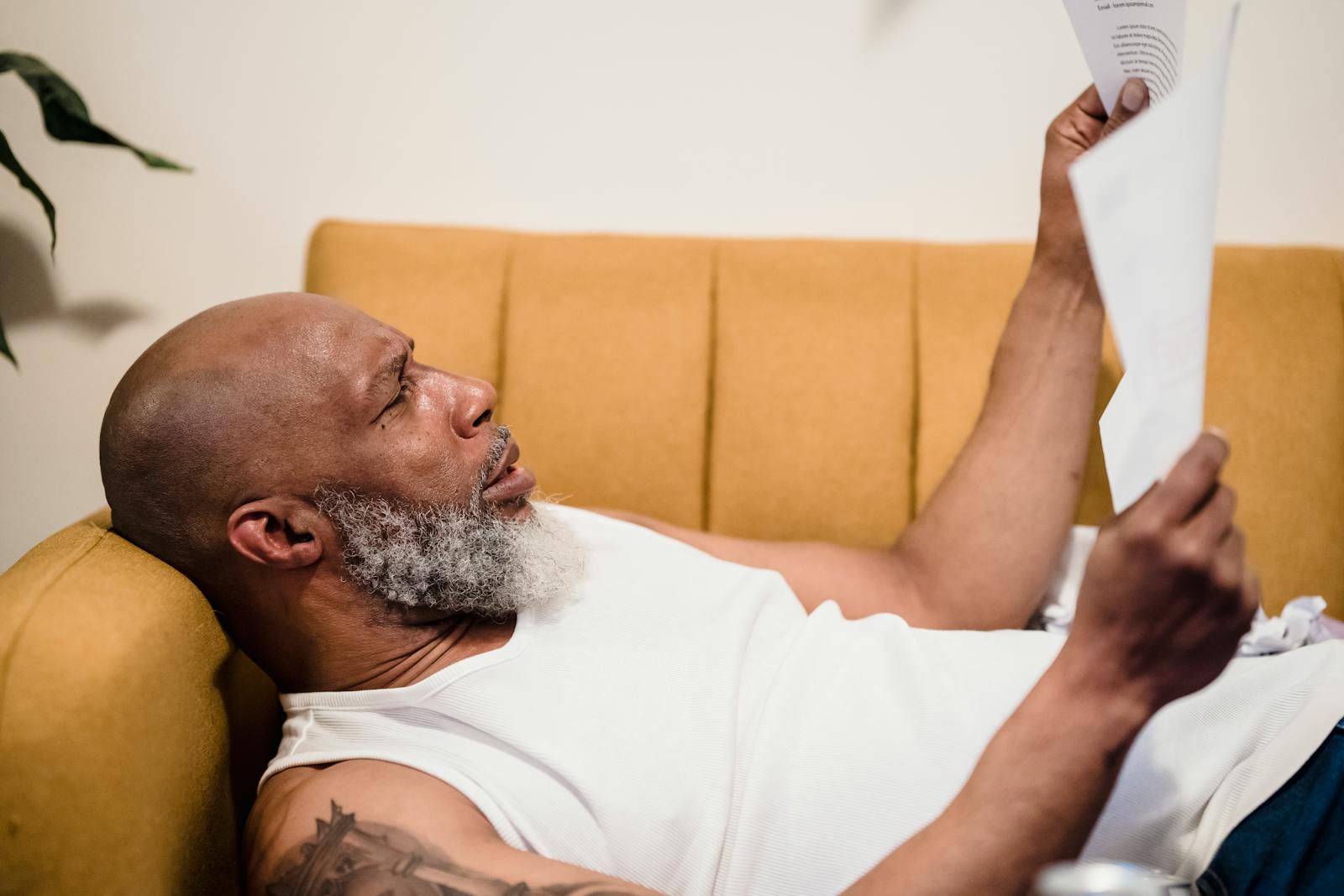 Bald man with facial hair lies on a mustard couch, reading documents in a relaxed setting.