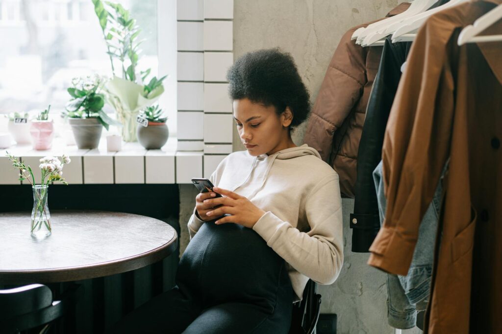 A pregnant woman with afro hair sits indoors, texting on her mobile phone by a window.