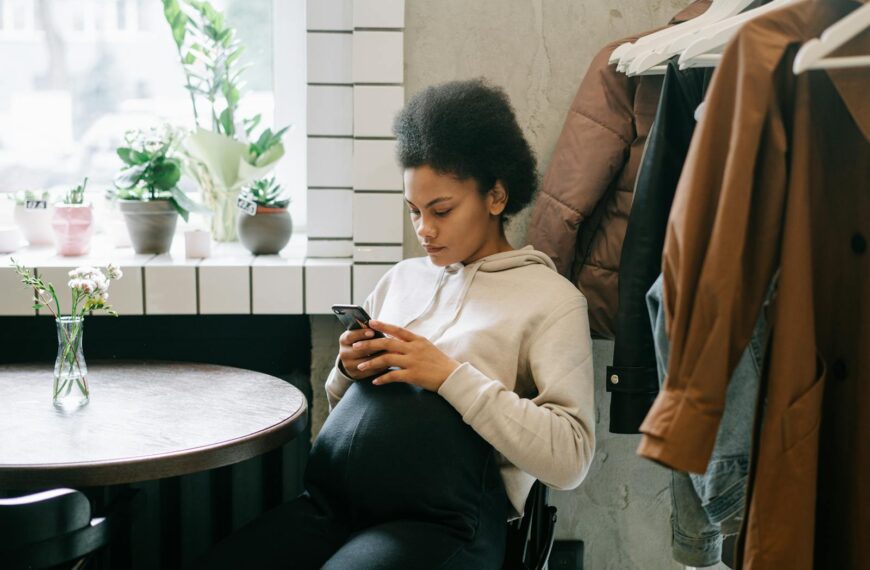 A pregnant woman with afro hair sits indoors, texting on her mobile phone by a window.