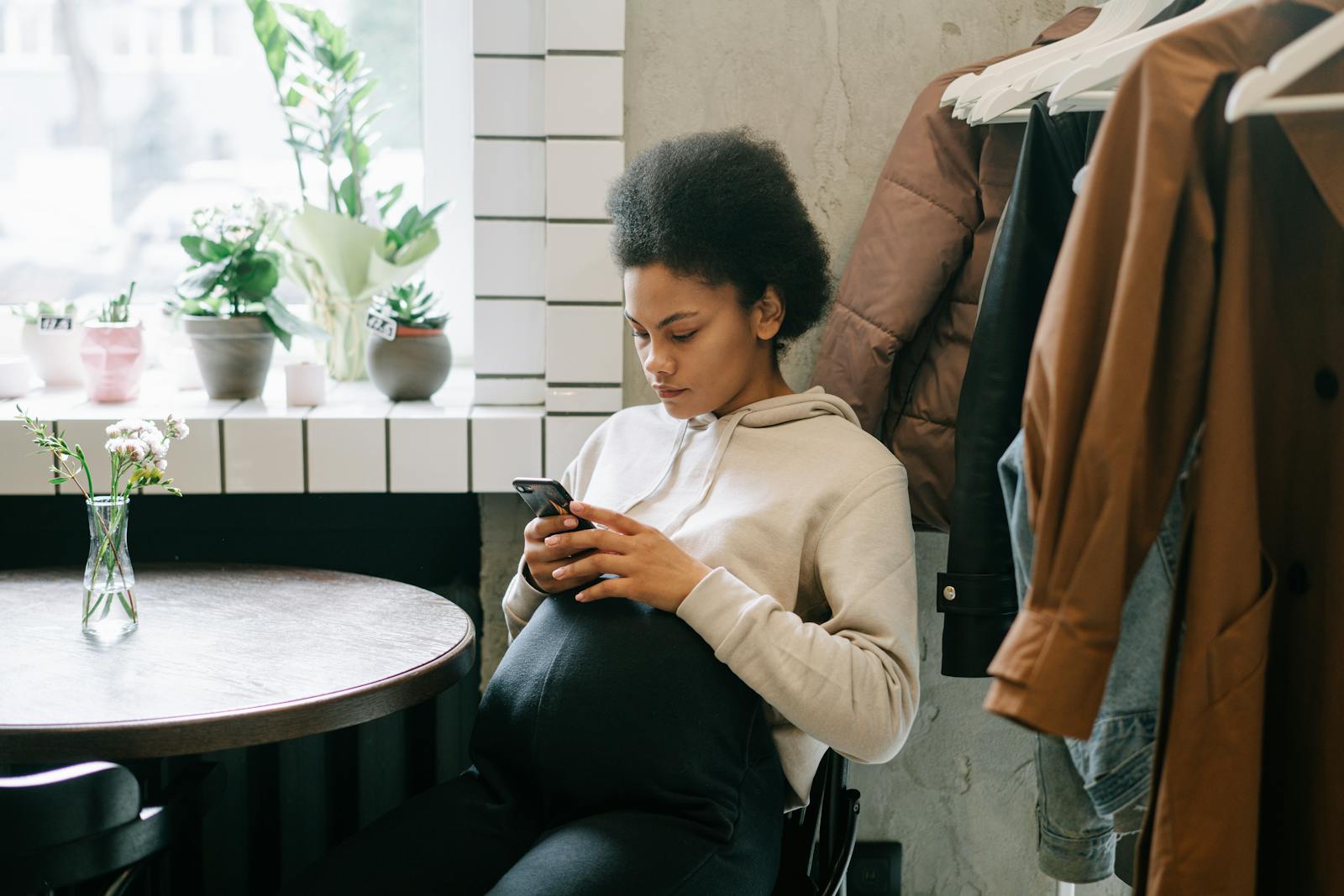 A pregnant woman with afro hair sits indoors, texting on her mobile phone by a window.