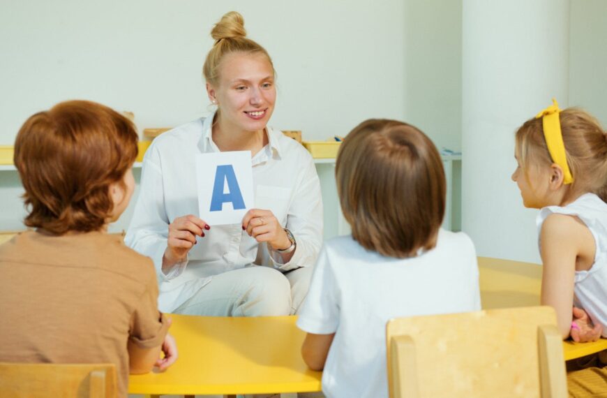 A teacher holds up a letter card while interacting with preschool children in a classroom setting.