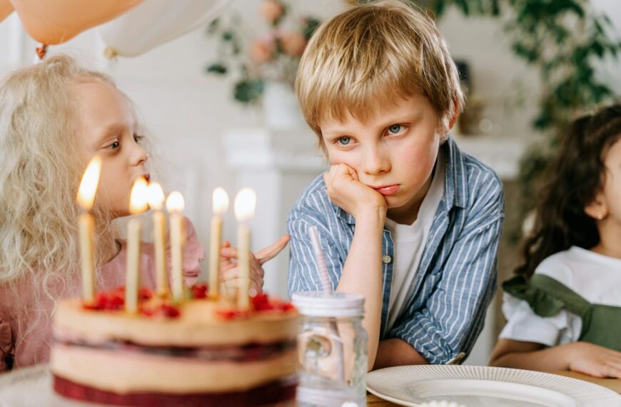 A young boy looking unhappy next to a birthday cake with lit candles, surrounded by friends.