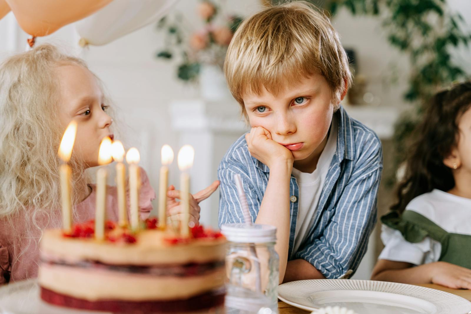 A young boy looking unhappy next to a birthday cake with lit candles, surrounded by friends.