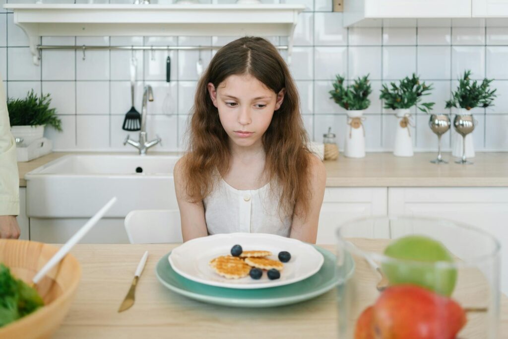A young girl looks down unhappily at her breakfast pancakes with blueberries in a modern kitchen.