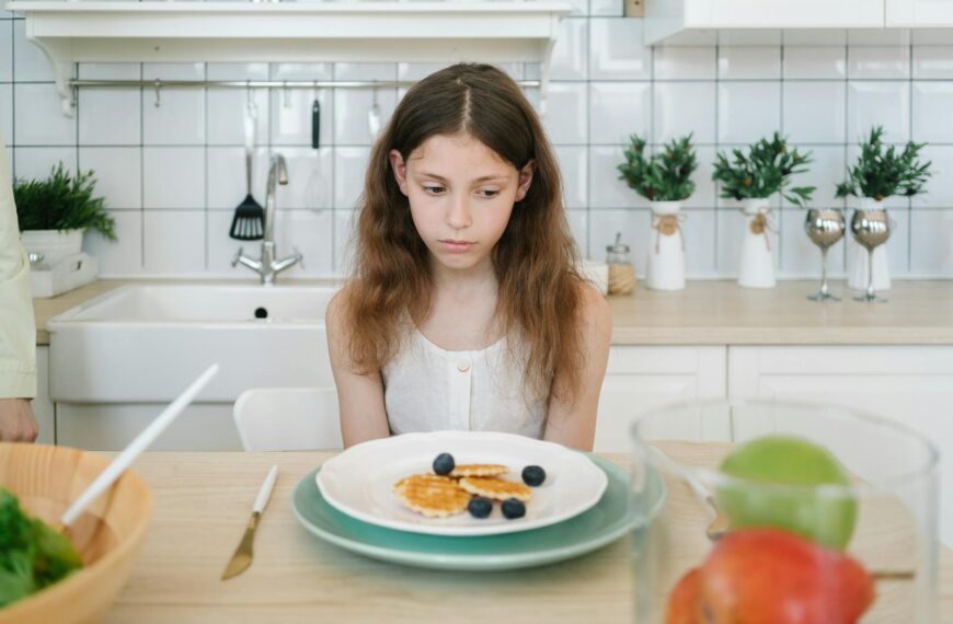 A young girl looks down unhappily at her breakfast pancakes with blueberries in a modern kitchen.