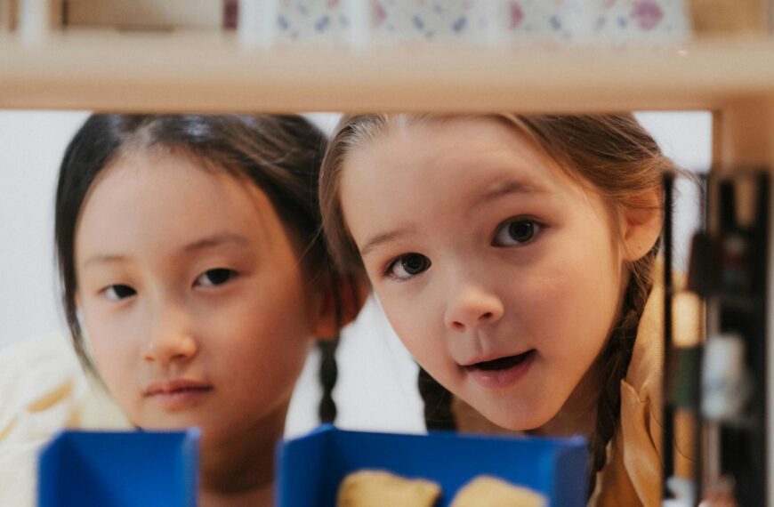 Two girls exploring with curious expressions, peering through shelves indoors.