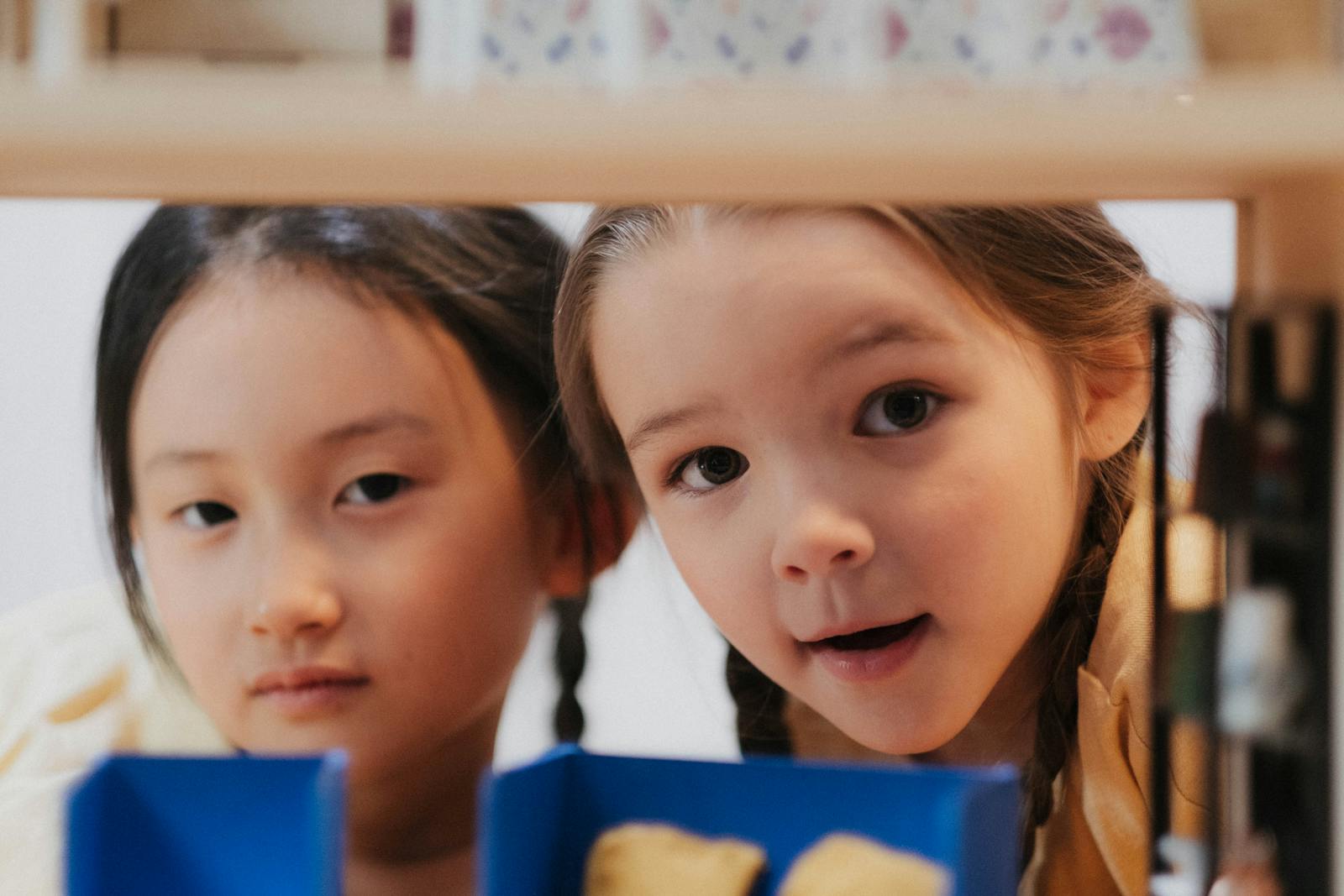 Two girls exploring with curious expressions, peering through shelves indoors.