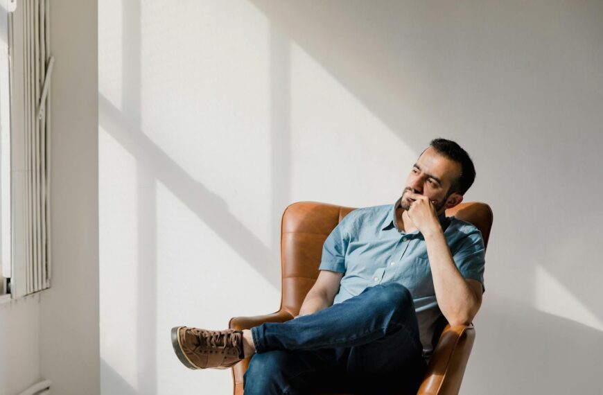 Man in casual attire sitting thoughtfully in a sunlit room, deep in contemplation.