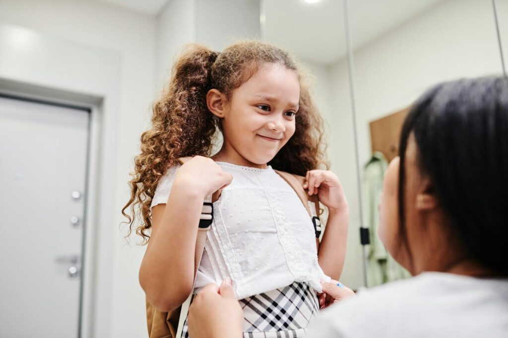 A smiling young girl with curly hair ready for school with her mother indoors.
