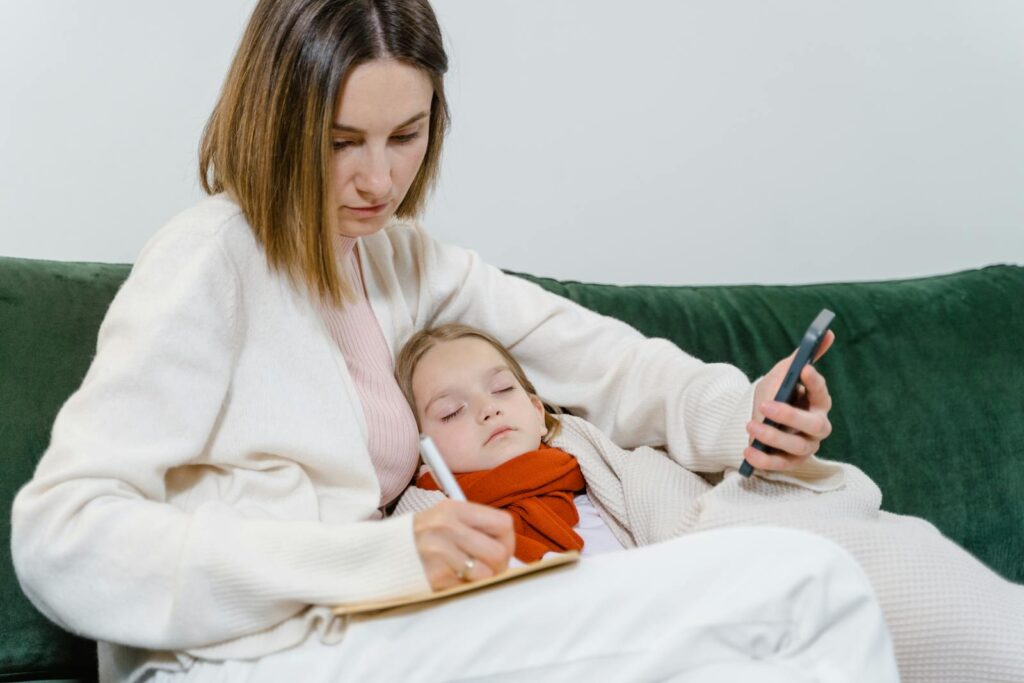 A mother comforts her sick child while writing notes and using a smartphone, portraying modern caregiving.