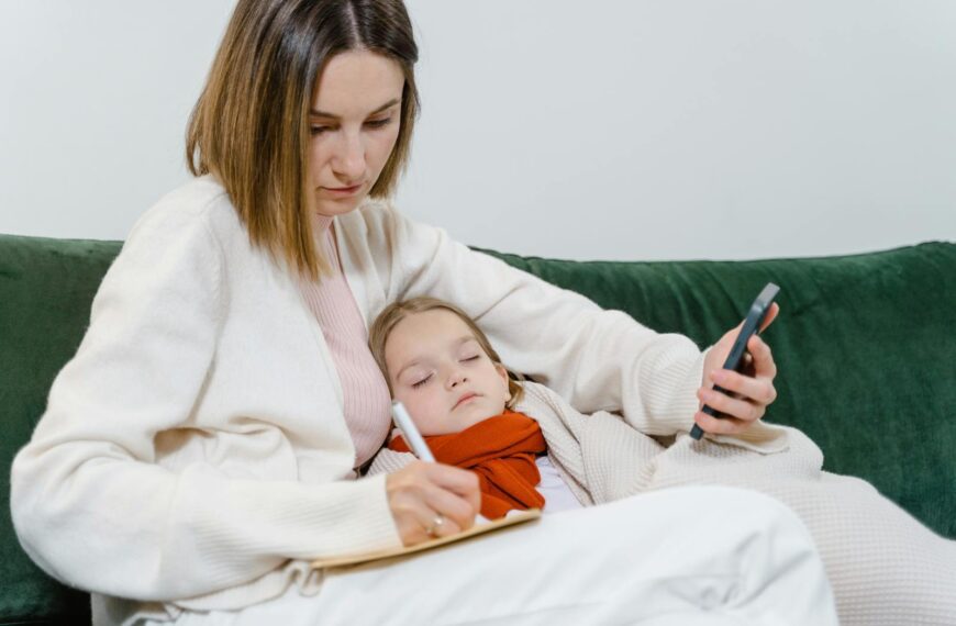 A mother comforts her sick child while writing notes and using a smartphone, portraying modern caregiving.