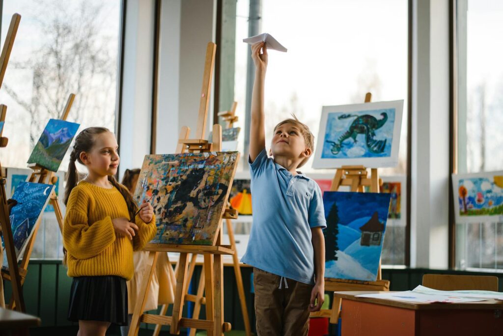 Kids enjoying paper planes amidst colorful paintings in a bright classroom.