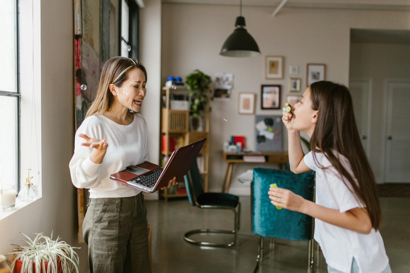 A mother works remotely while her daughter plays with bubbles in a cozy home setting.