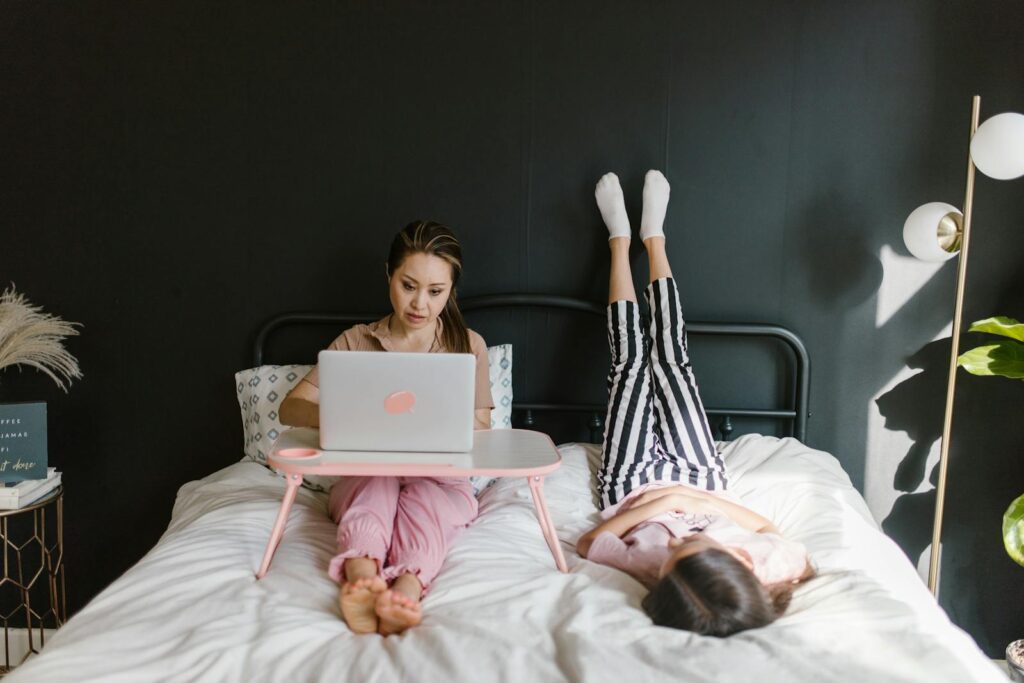 Woman working remotely on laptop in bed, child playing nearby, embracing new work-from-home culture.