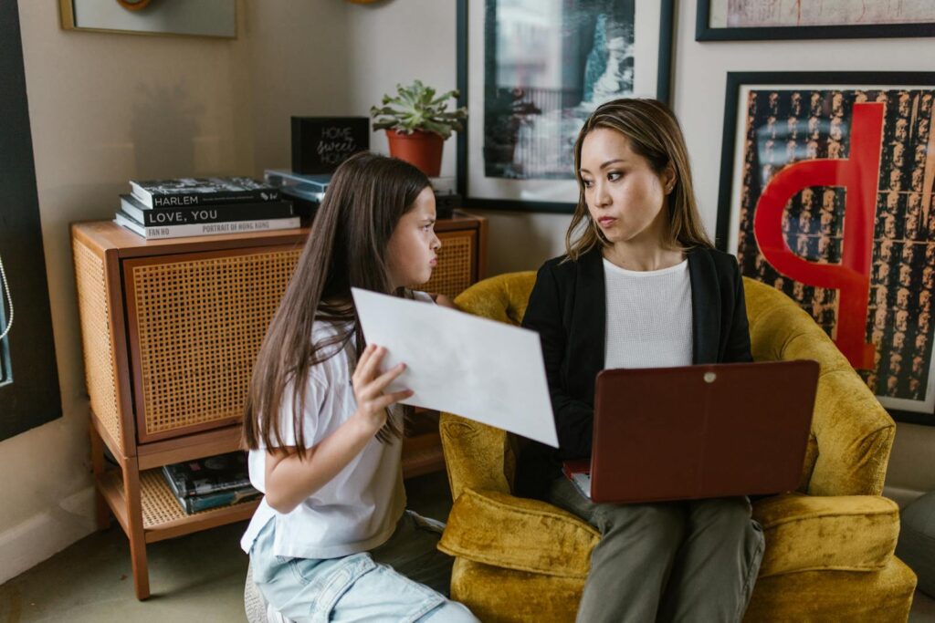 An Asian woman and her daughter collaborating at home, using a laptop and paper.