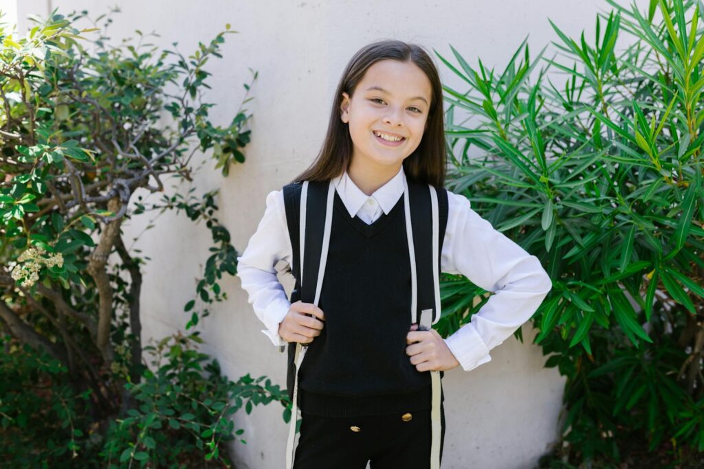 Young girl in school uniform smiling confidently outdoors with backpack.