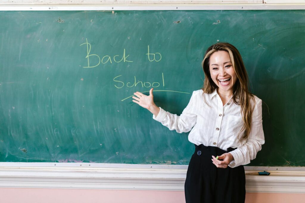 Smiling teacher in a classroom pointing at back to school message on a chalkboard.