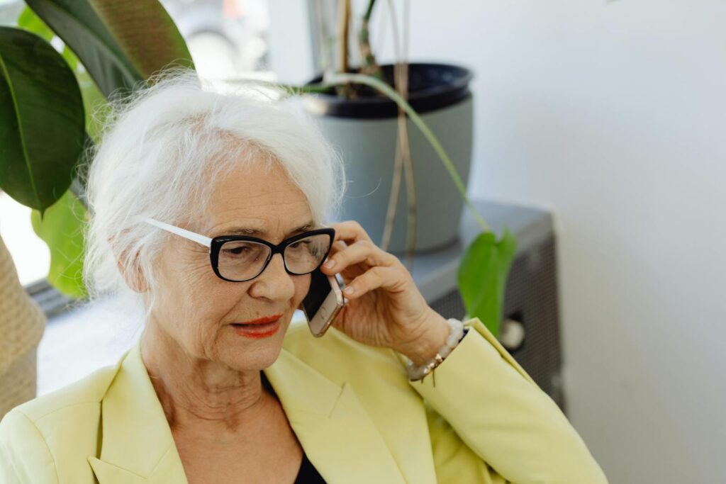 Elderly woman with white hair and eyeglasses using cellphone indoors.