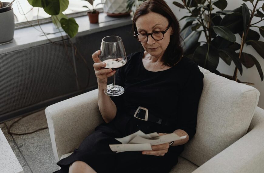 Elderly woman seated on a sofa holding a glass of wine, reflecting in a cozy room.