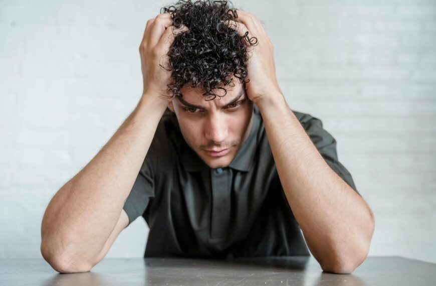 A young man sits indoors with his head in his hands, showing stress and worry.