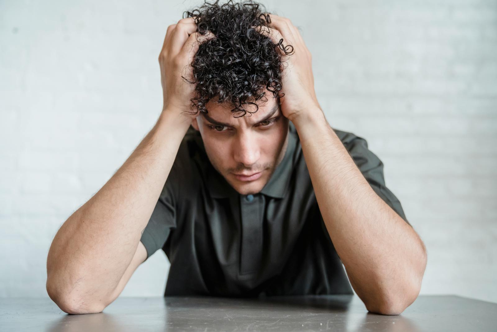 A young man sits indoors with his head in his hands, showing stress and worry.