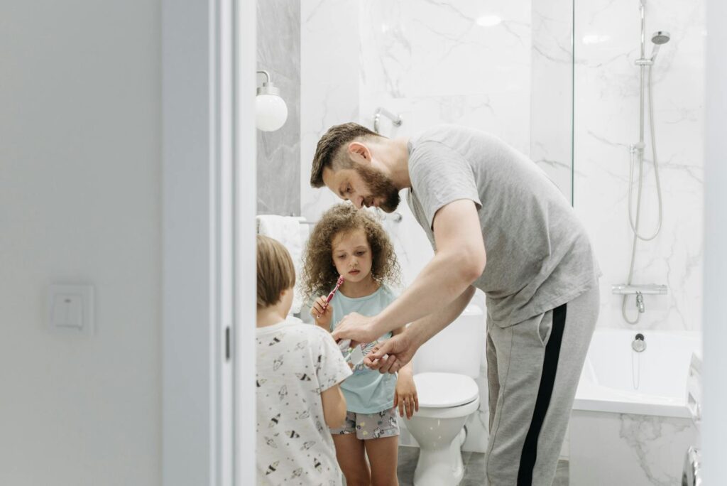 A father assists his children with brushing their teeth in a modern bathroom.