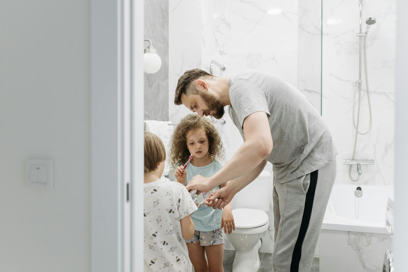 A father assists his children with brushing their teeth in a modern bathroom.