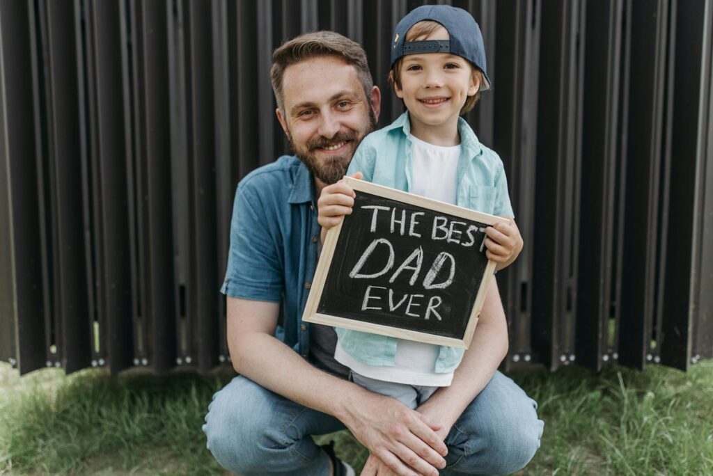 A father and son smiling together holding a 'Best Dad Ever' sign outside.