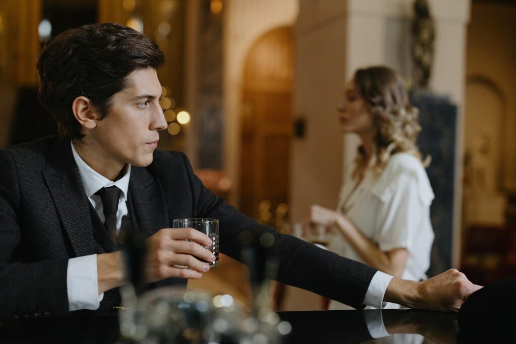 A sophisticated man in a suit holds a drink at a chic bar with a woman in the background.
