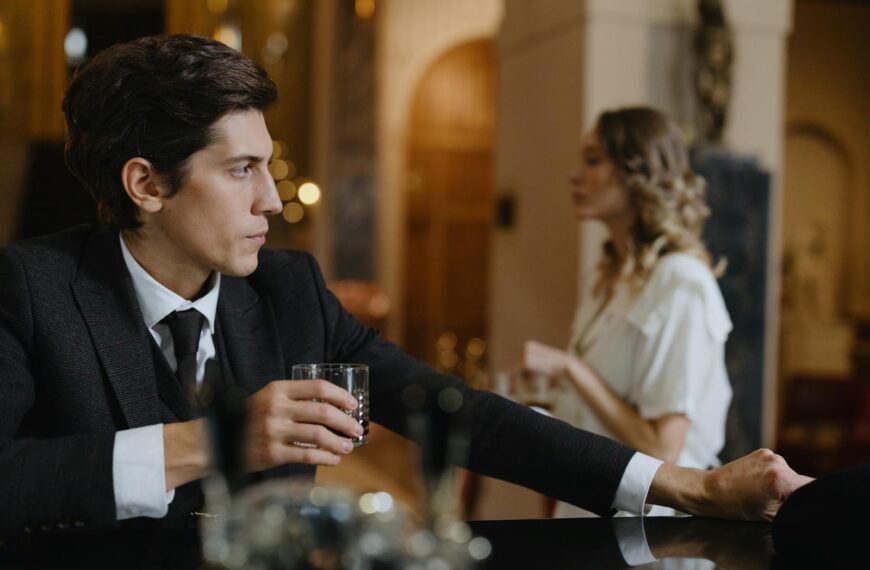 A sophisticated man in a suit holds a drink at a chic bar with a woman in the background.